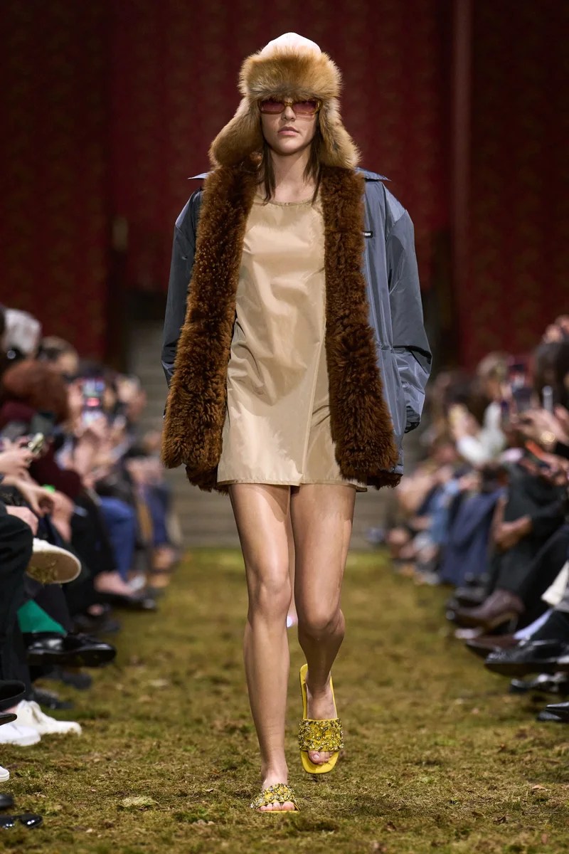 Модель Miu Miu у куртці з хутряною підкладкою поверх короткої бежевої сукні і в хутряній шапці-вушанці